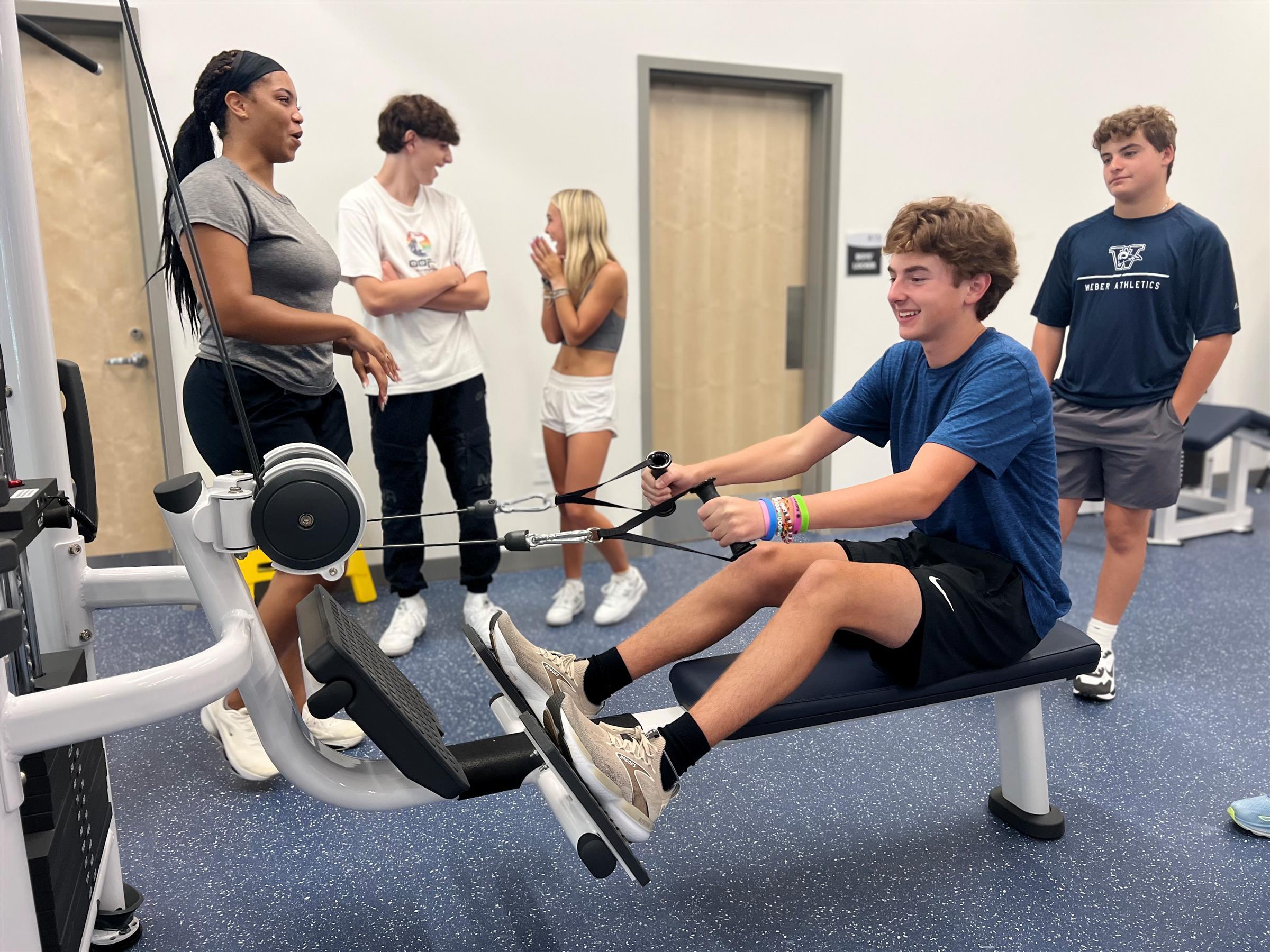 students in a gym setting, with one using a rowing machine, smiling. 